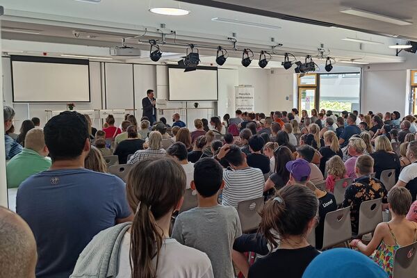 Der Schulleiter begrüßt die neuen Schülerinnen und Schüler mit Eltern in der Mensa.